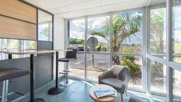 A modern hotel room with large windows, a seating area with a table, and a view of the street and greenery outside.
