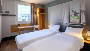Habitación de hotel con dos camas individuales, un escritorio, una silla, un armario y un ventanal con vista a un edificio.
