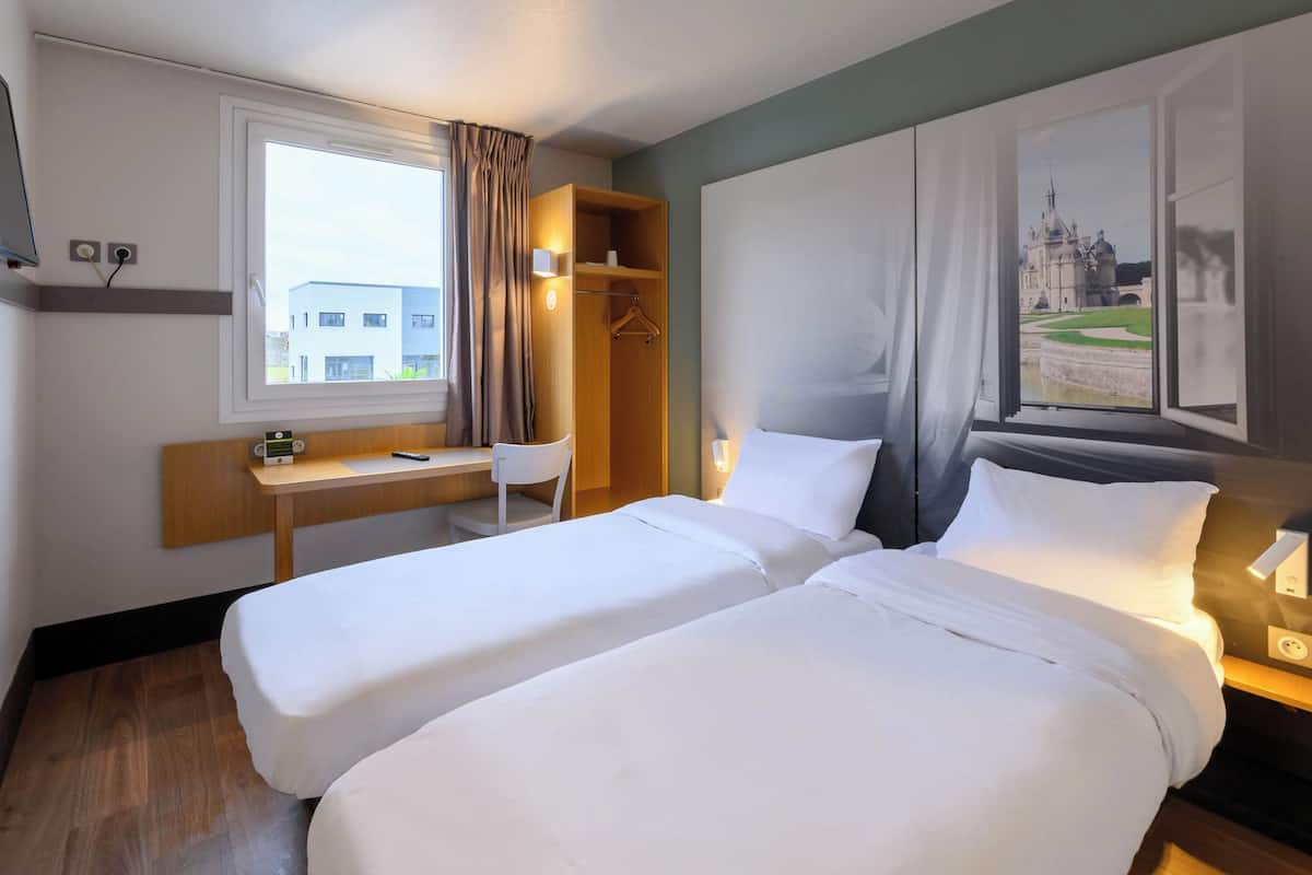 Hotelkamer met twee enkele bedden, een bureau, een stoel, een kledingkast en een groot raam met uitzicht op een gebouw.