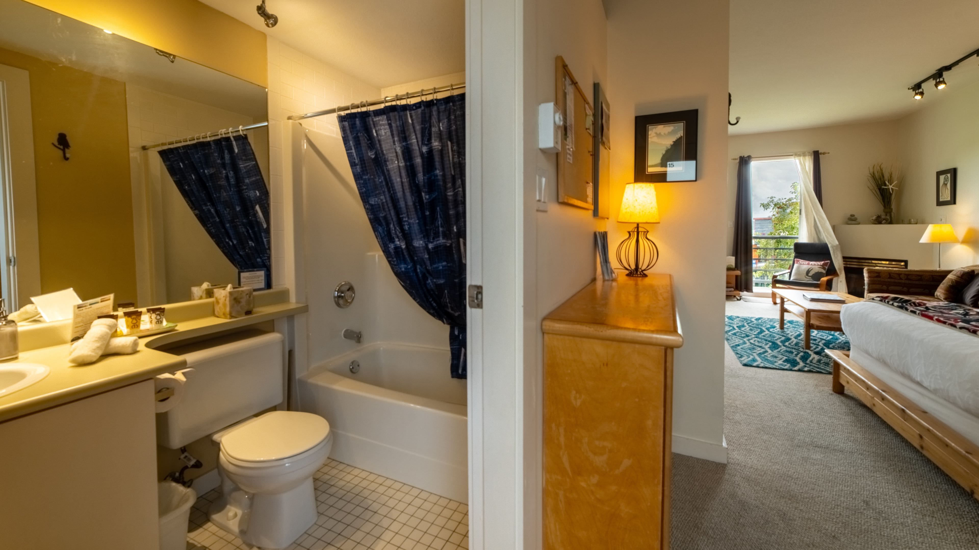 Une salle de bain avec une baignoire, des toilettes et un lavabo. Il y a un rideau de douche et un miroir au-dessus du lavabo. La pièce est dotée d’un plancher en bois et d’une fenêtre donnant sur l’extérieur.
