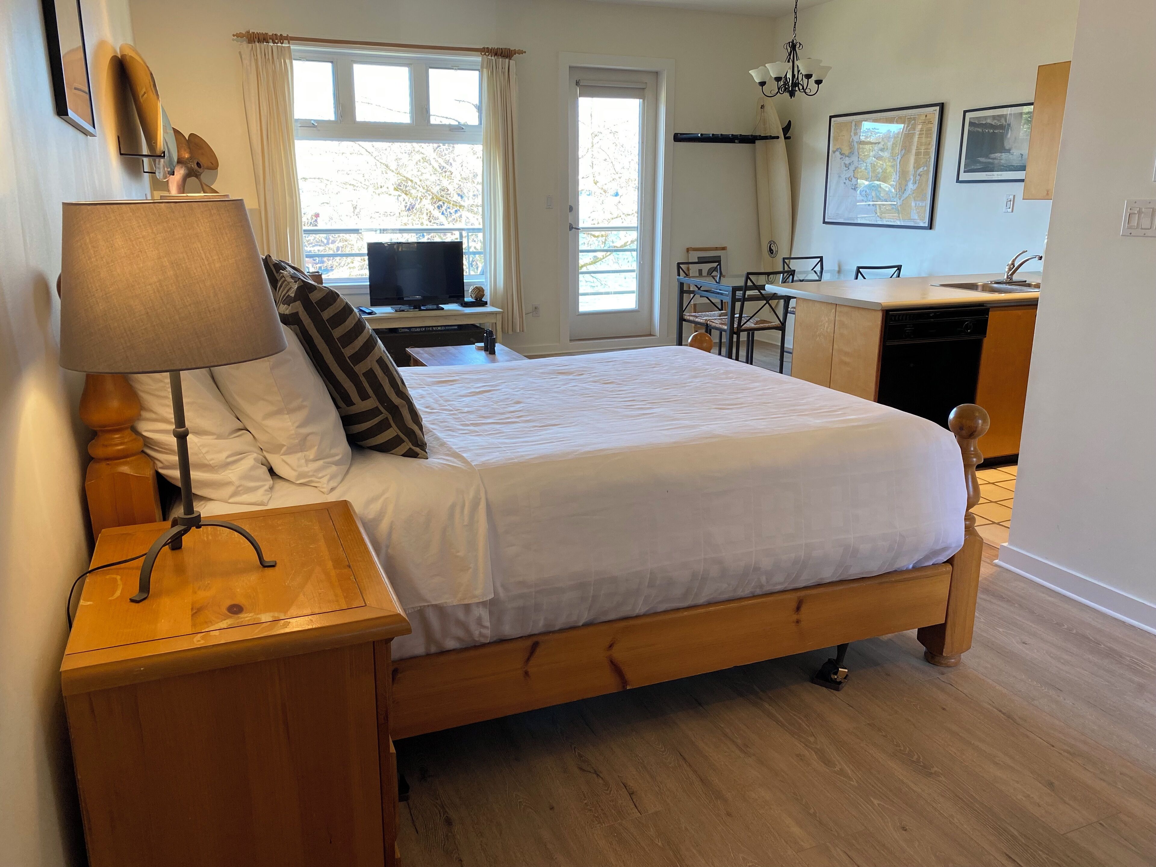 Un dormitorio con cama de madera, una mesita de noche con lámpara, un escritorio con computadora, una silla y vistas al exterior a través de una ventana.