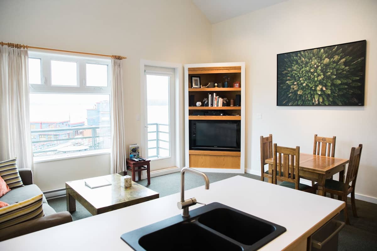 A modern kitchen with a sink, dining table, and chairs. There is a bookshelf with books and a television.