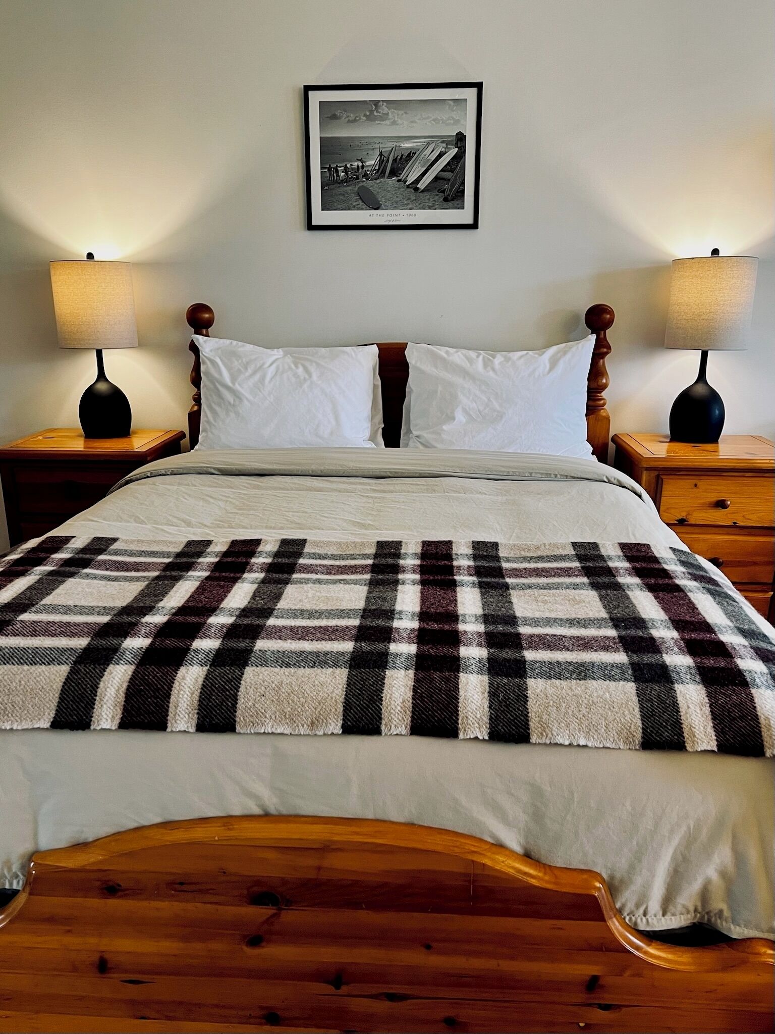 Un lit bien fait, recouvert d’une couverture à carreaux, deux tables de chevet avec des lampes, un tableau encadré au mur et une tête de lit en bois.