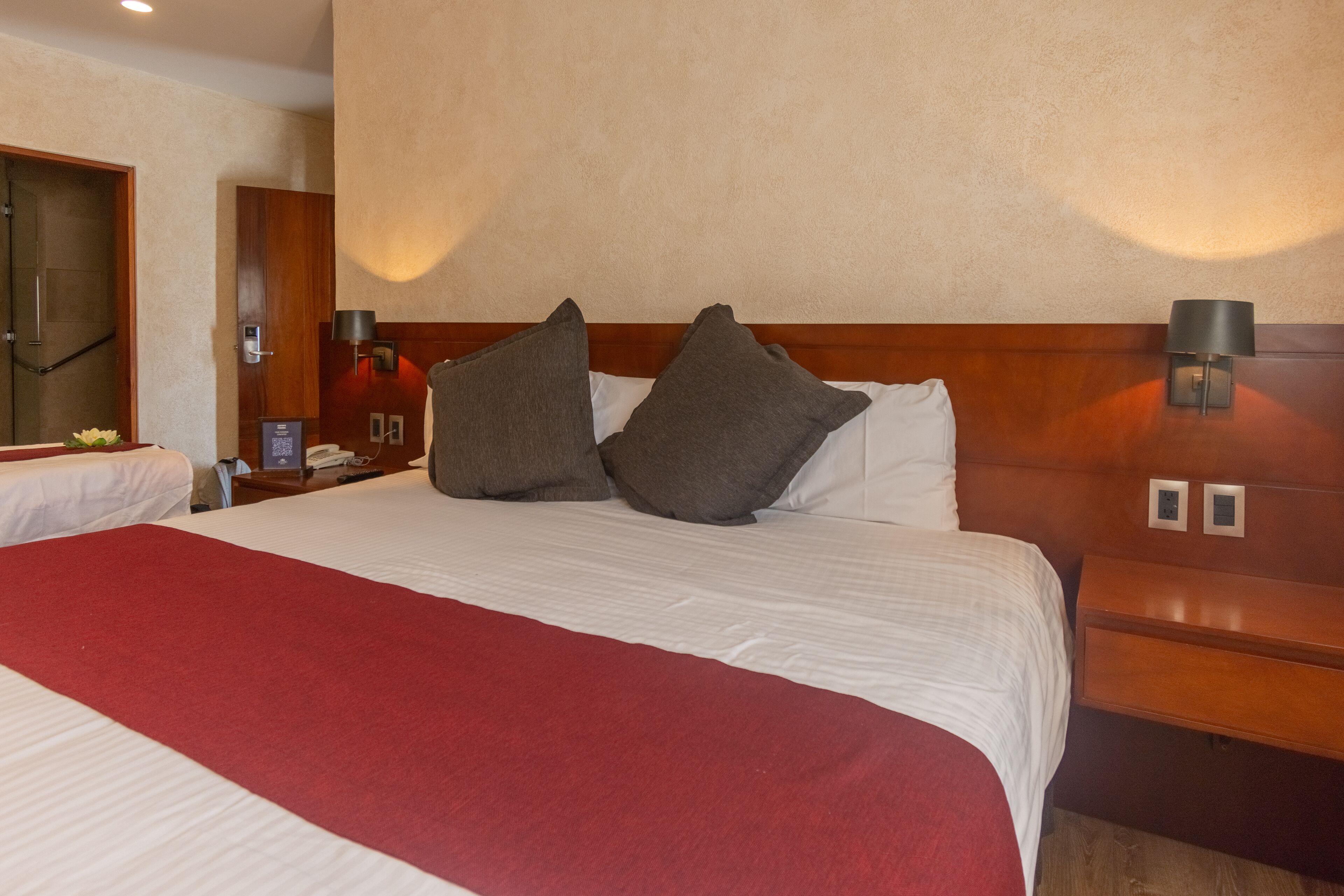 A hotel room with a large bed, two bedside tables, a nightstand, a mirror, and a red throw blanket.