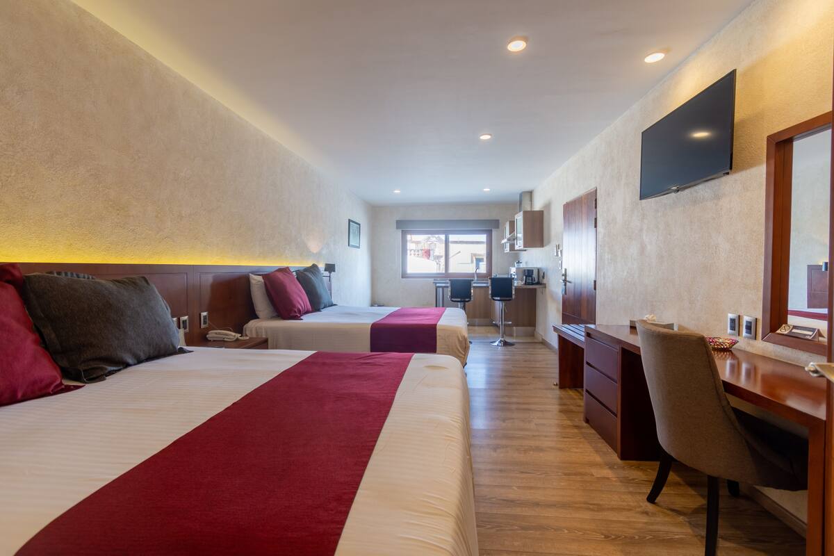 A hotel room with two beds, a desk, a chair, a TV, and a small kitchenette.