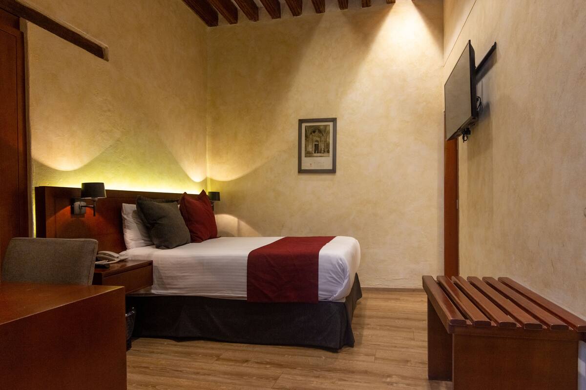 A hotel room with a bed, bedside table, lamp, and a bench.