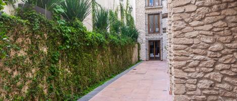 A narrow pathway flanked by lush greenery and a stone wall, leading to a building with a stone facade and multiple windows.