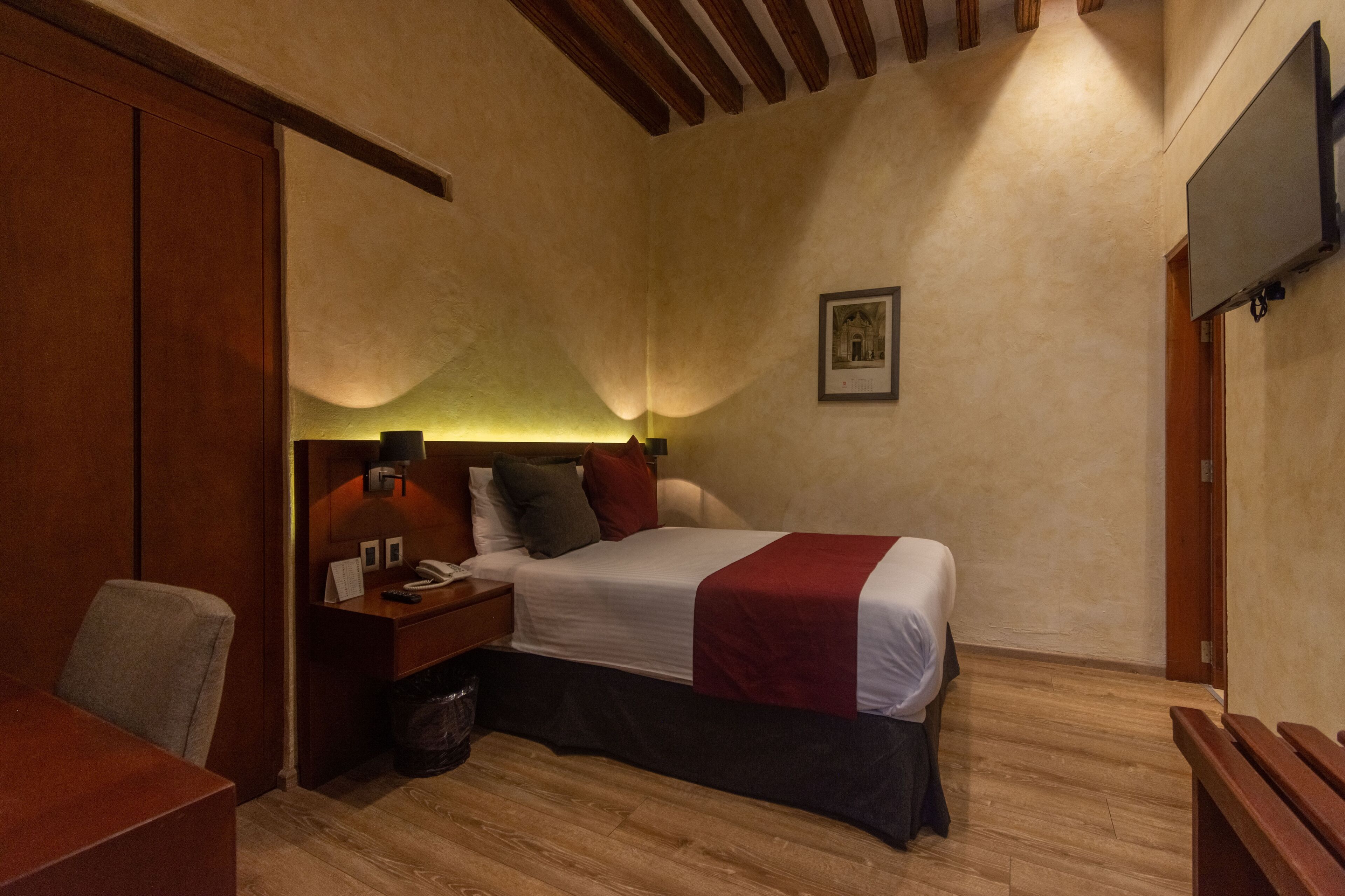 A hotel room with a bed, bedside table, lamp, and a television mounted on the wall.