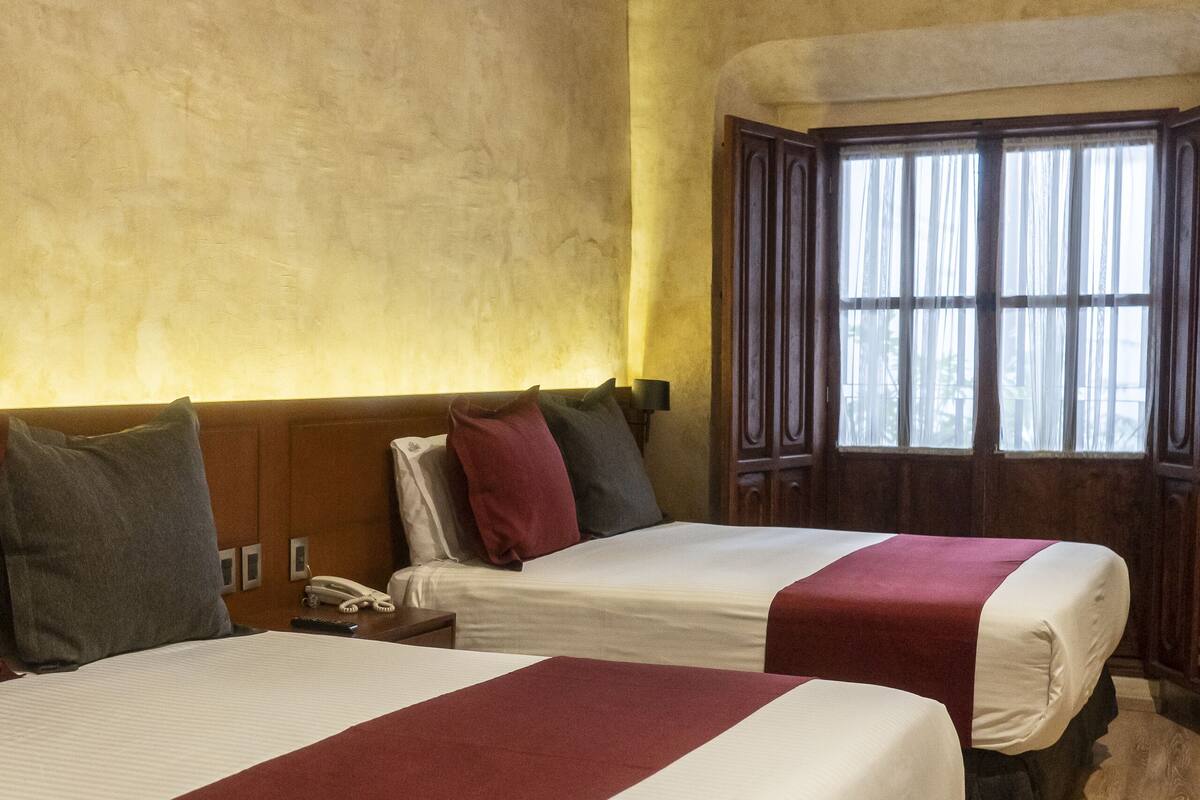 A hotel room with two beds, wooden flooring, a window with curtains, and a wooden ceiling.