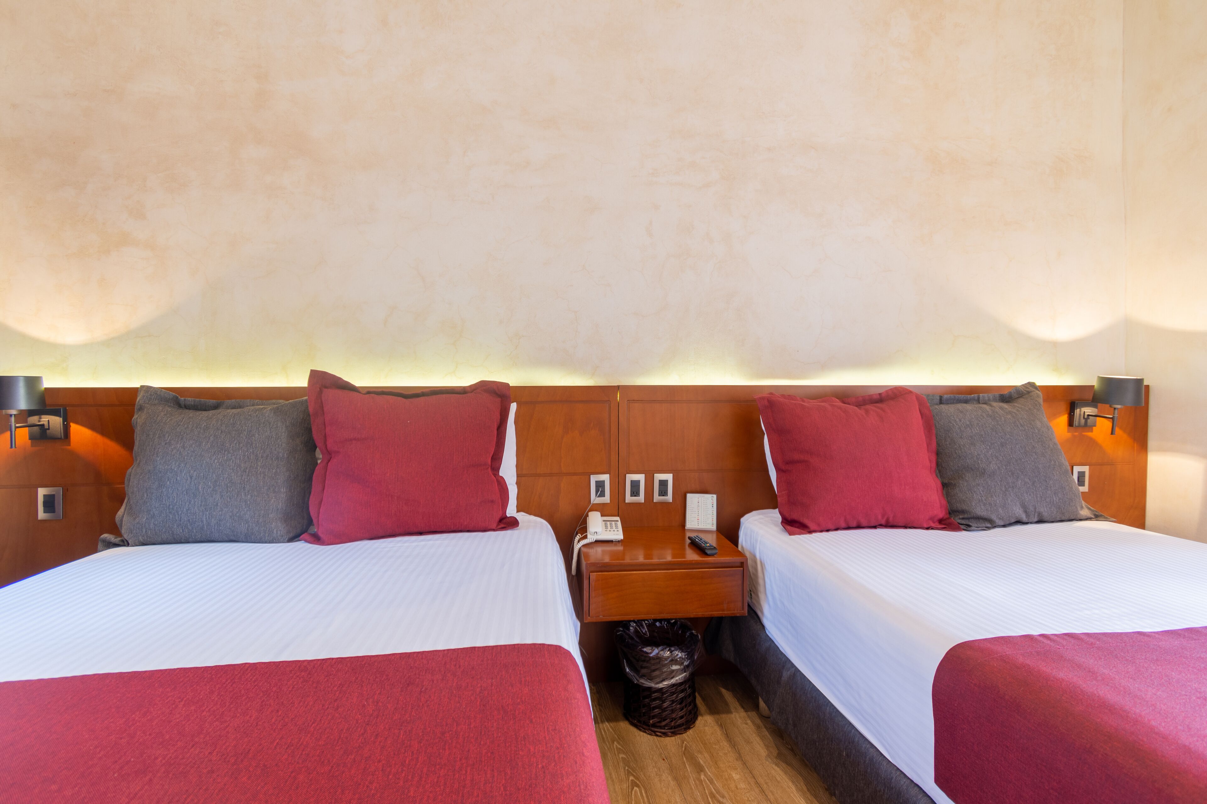 Habitación de hotel con dos camas, cada una con cabecera de madera, mesita de noche y cubo de basura.