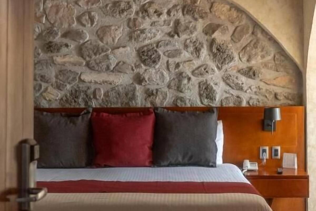 A bedroom with a stone wall, a wooden headboard, a bed with a red pillow, and a nightstand with a lamp.