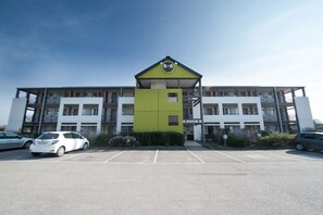 Un edificio de varios pisos con fachada verde y blanca, un estacionamiento al frente y un letrero que indica un espacio para negocios u oficinas.