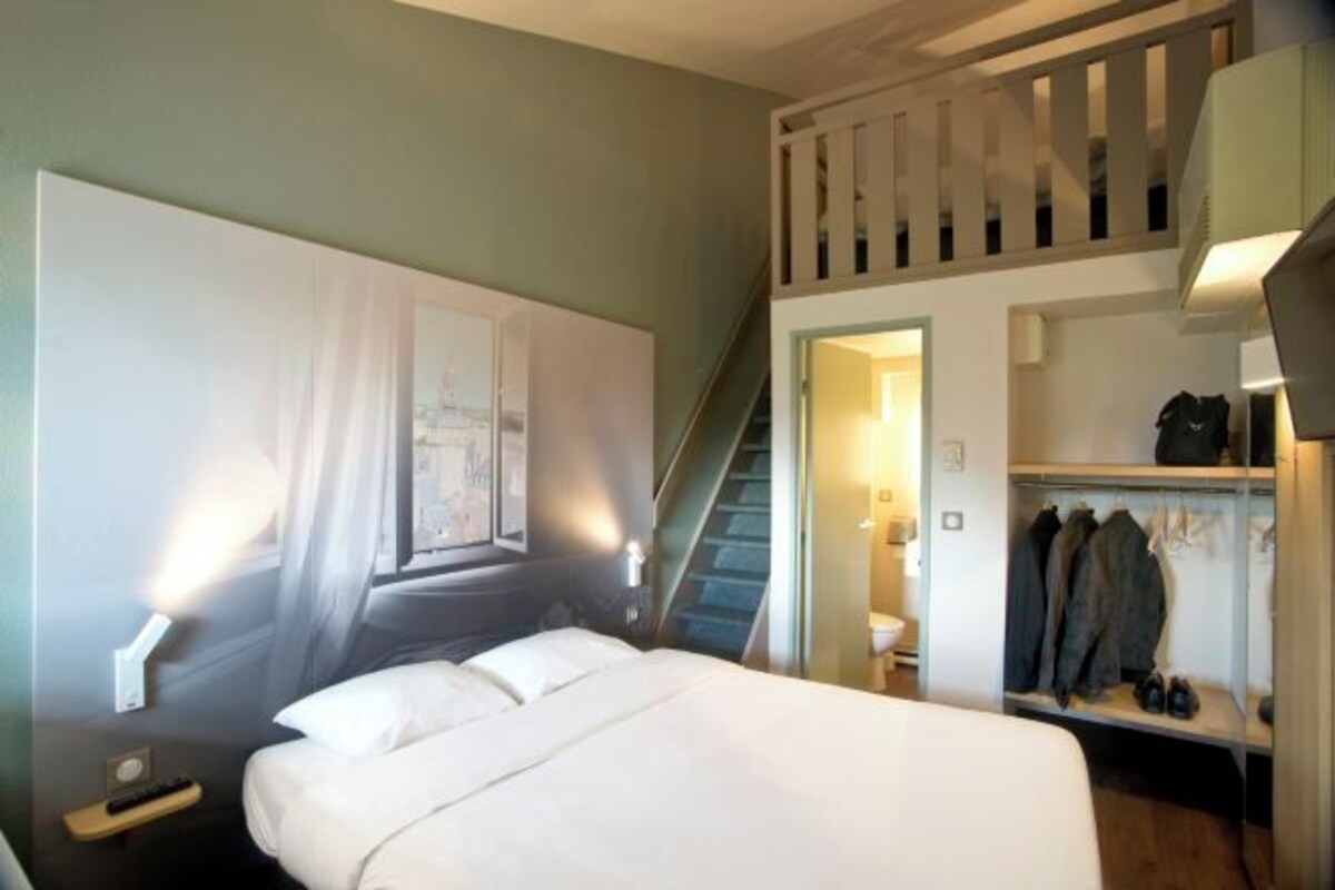 Habitación de hotel con cama, mesita de noche, lámpara de pared, perchero con abrigos y una escalera que sube a un nivel superior.