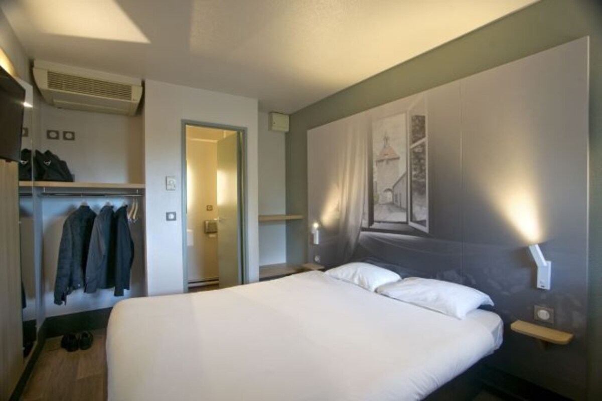 Una habitación de hotel con una cama, un armario, perchero y una puerta que da a otra habitación.