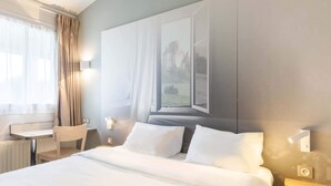 A hotel room with a large bed, a desk, and a chair. There is a window with a view of a castle. - B&B HOTEL Moulins Toulon‑sur‑Allier (Toulon-sur-Allier)