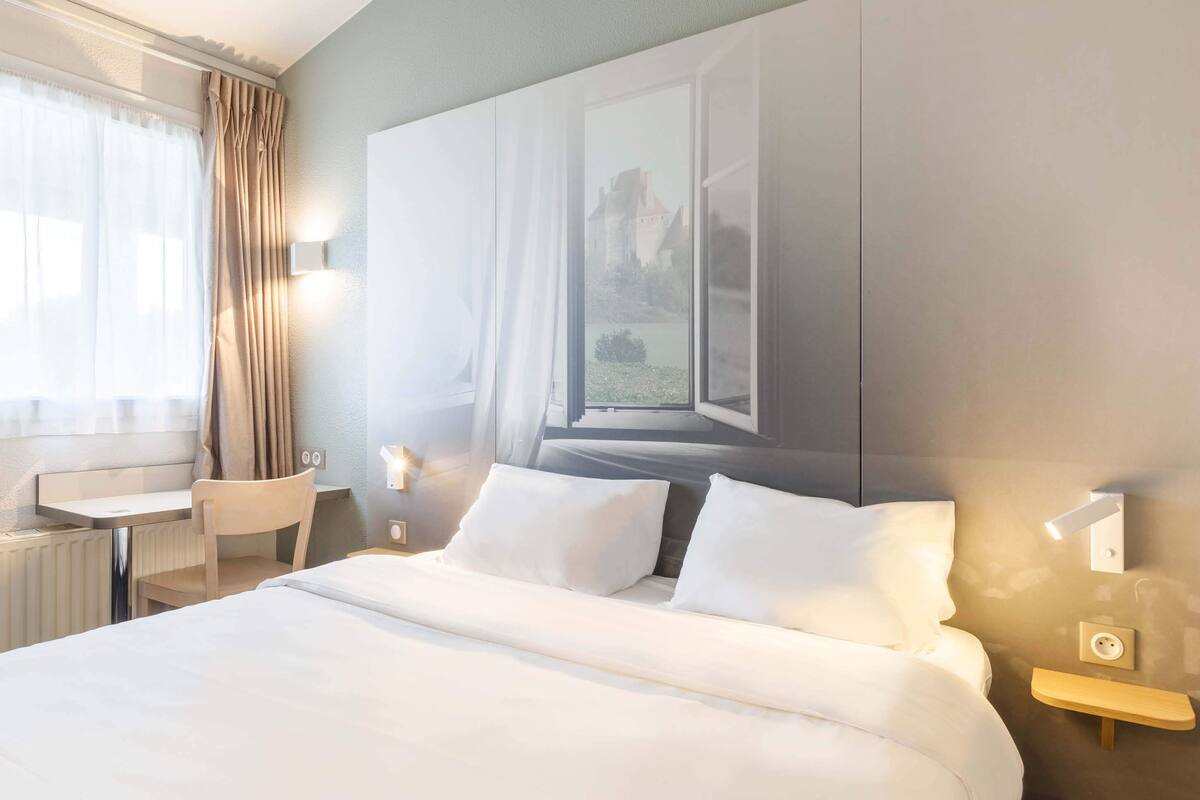 A hotel room with a large bed, a desk, and a chair. There is a window with a view of a castle.