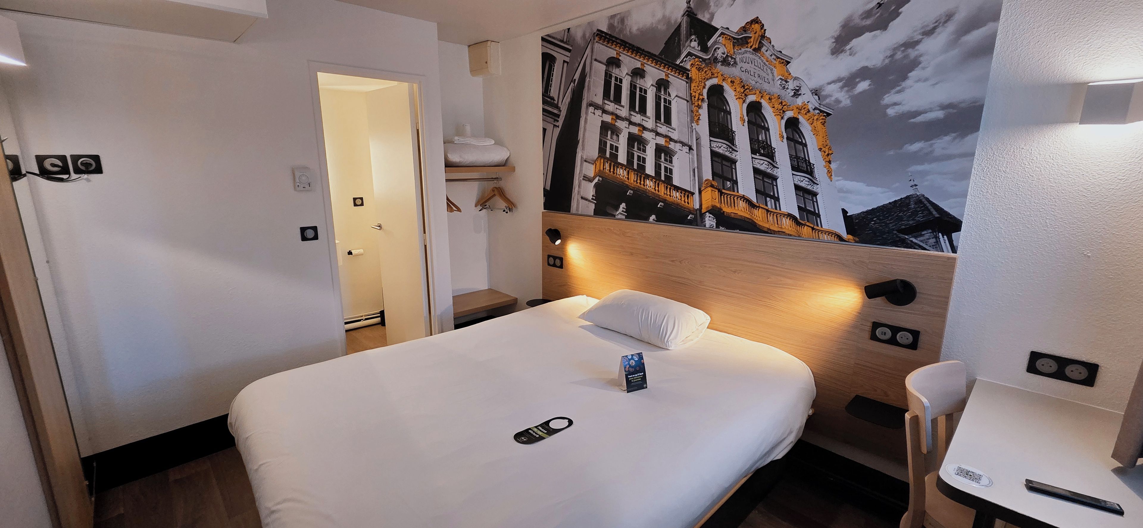 A hotel room with a bed, a bedside table, a chair, a wall-mounted light, and a door.