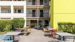 An outdoor seating area with tables and chairs, a building with balconies, and a yellow wall. - B&B HOTEL Moulins Toulon‑sur‑Allier (Toulon-sur-Allier)