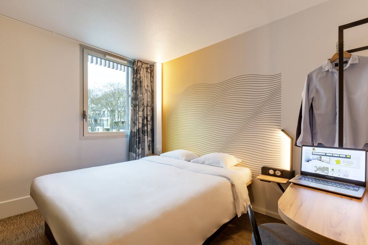 Une chambre d’hôtel avec un lit, un ordinateur portable posé sur une table de chevet et une fenêtre avec des rideaux.