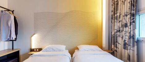 A hotel room with two beds, a bedside table, a lamp, a wardrobe, and a window with curtains.