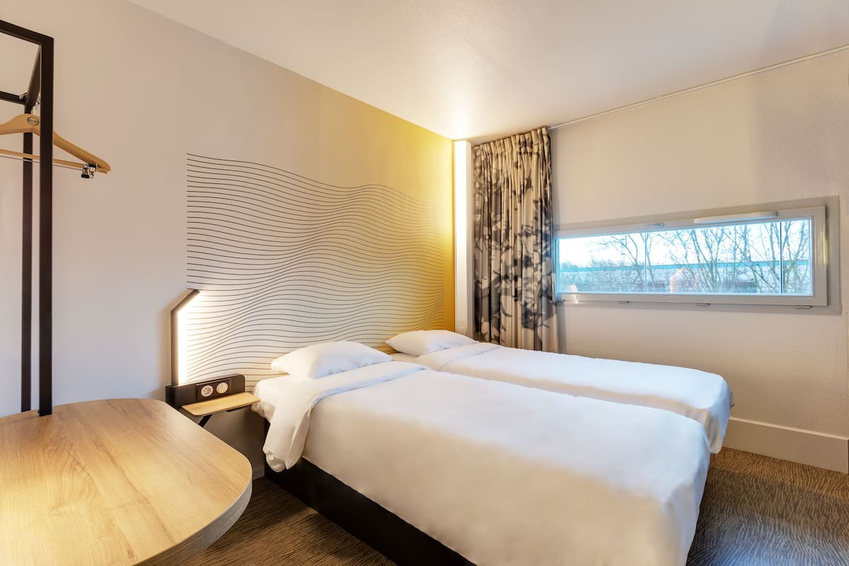 Une chambre d’hôtel avec deux lits, une table en bois, une lampe fixée au mur et une fenêtre avec des rideaux.