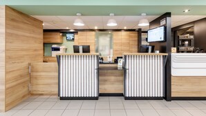 Zona de recepción con paneles de madera, una mampara de vidrio y una pantalla digital que muestra información.