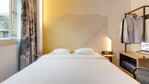 Habitación de hotel con cama, escritorio con laptop, silla, perchero y ventana con cortinas.