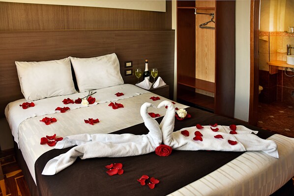 A hotel room with a neatly made bed, a wooden headboard, a nightstand with a bottle and glasses, and a bathroom visible through an open door.