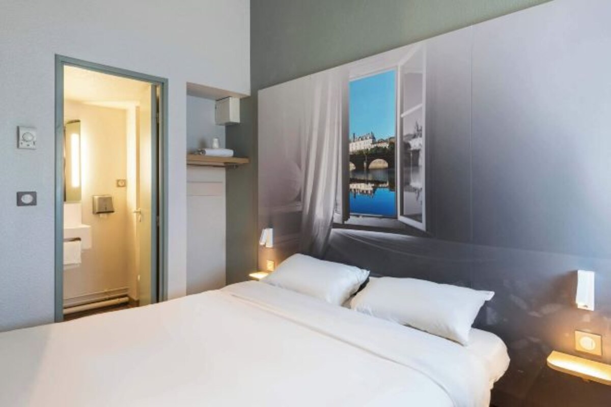A hotel room with a bed, a nightstand with a lamp, a wall-mounted shelf, and a door leading to a bathroom.