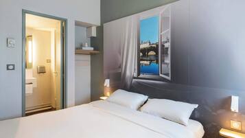 Et hotelværelse med en seng, et badeværelse der kan ses igennem en åben dør, en vægmonteret lampe og et stort vindue med udsigt over bybilledet.