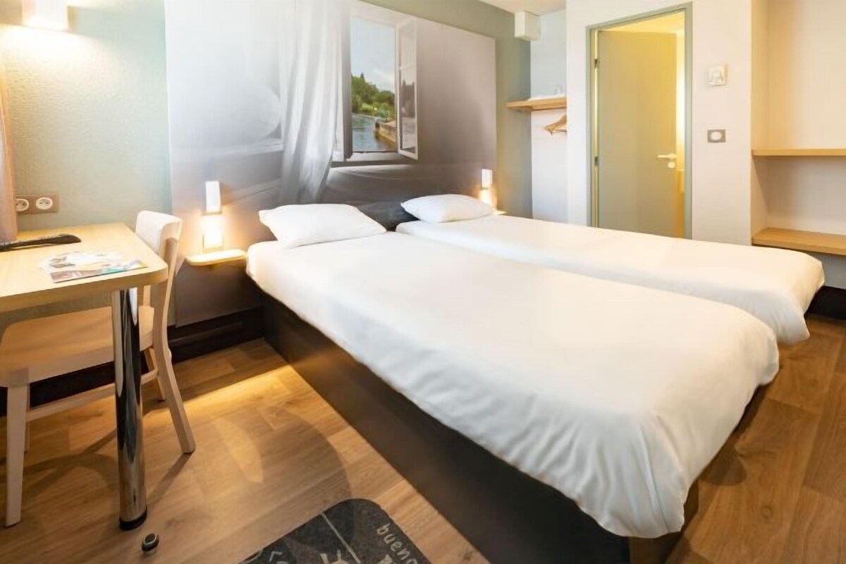 A hotel room with two beds, a desk, a chair, and a window with a view of greenery.