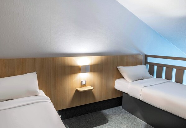 A small, cozy room with a sloped ceiling, wooden paneling, a bedside table with a lamp, and two beds.