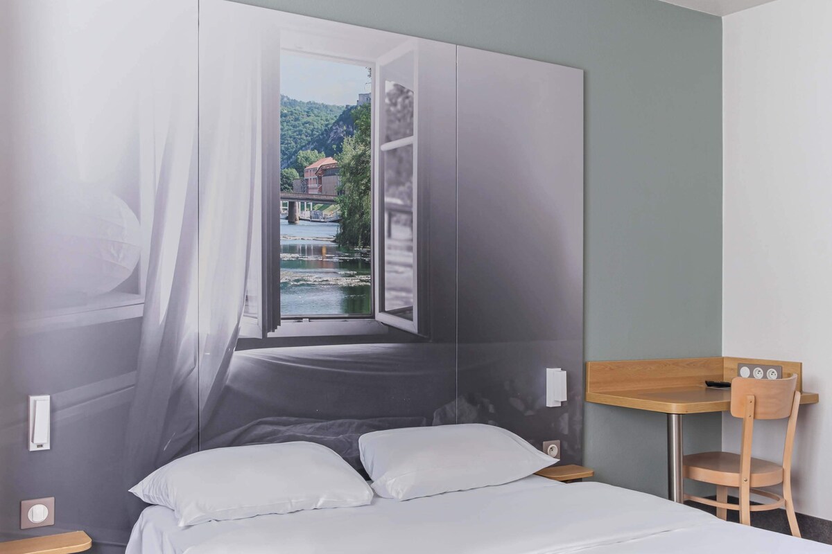A bedroom with a bed, a wooden chair, a small table, and a window with a view of a river and buildings.
