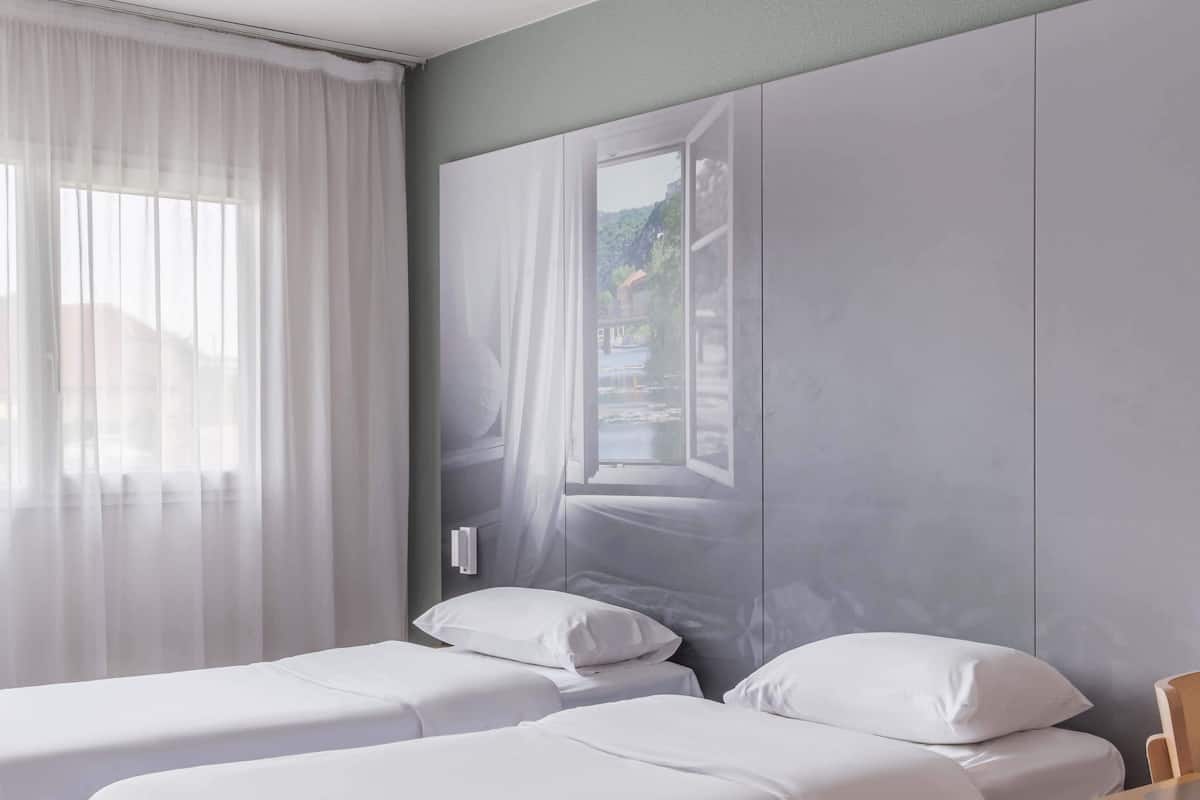 A bedroom with two beds, a large wardrobe, and a window with sheer curtains.