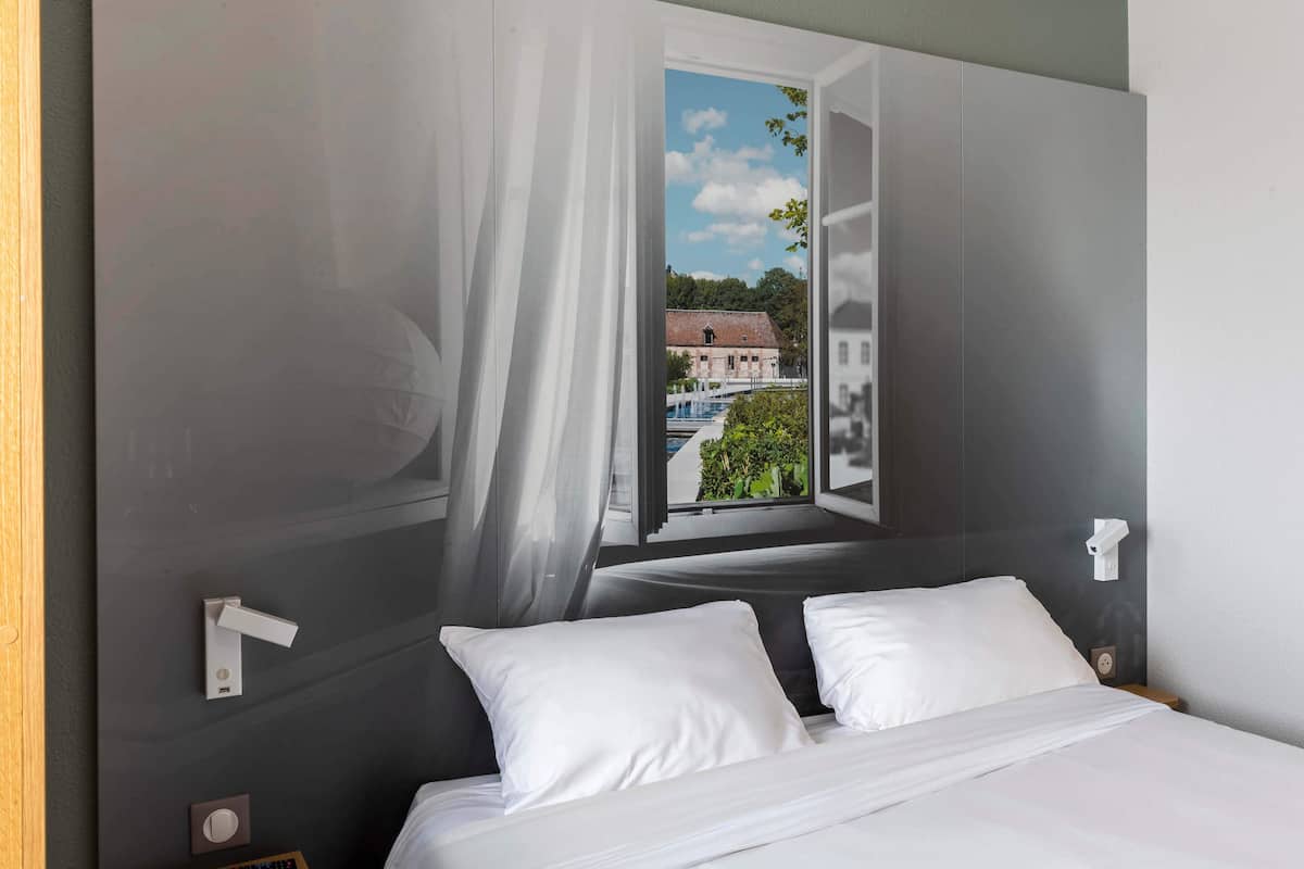 Ein Schlafzimmer mit einem Bett, weißen Kissen, einem Fenster mit Vorhängen und Blick auf ein Gebäude und Bäume.