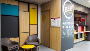 A B&B hotel reception area with a wooden panel wall, a yellow table, two leather armchairs, and a reception desk.