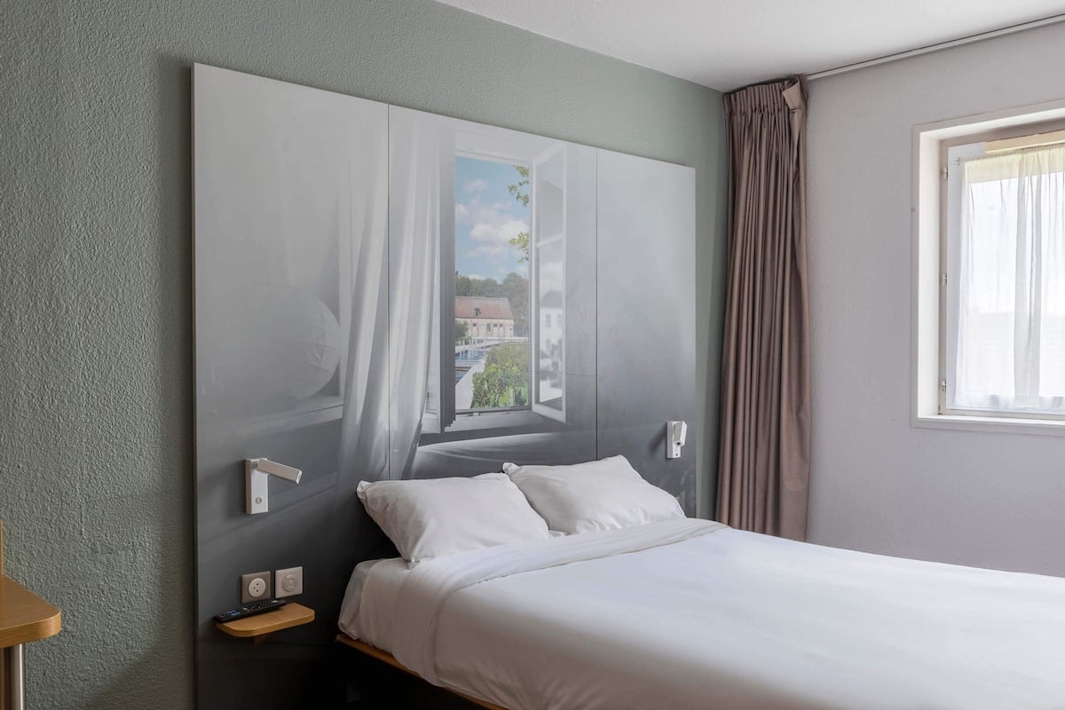 Ein Hotelzimmer mit einem großen Bett, Blick auf ein Gebäude durch ein Fenster und einem Vorhang.