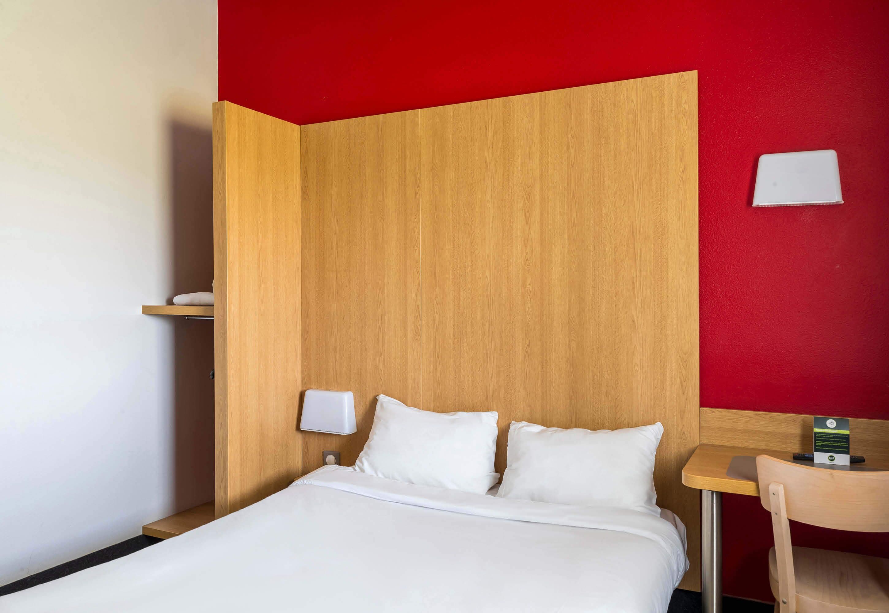 A hotel room with a wooden headboard, a white bed, a small table with a chair, and a red accent wall.