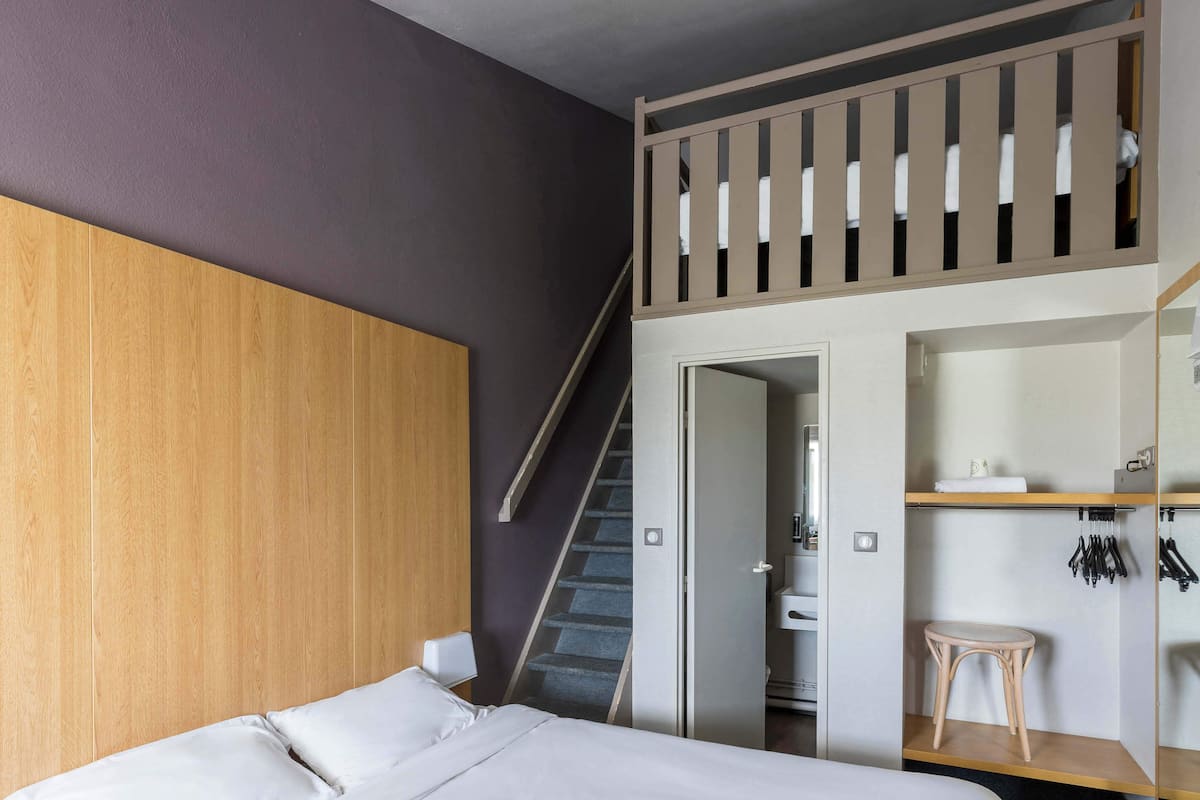 Une chambre avec un lit superposé, une tête de lit en bois, un escalier, une salle de bain et un petit tabouret en bois.