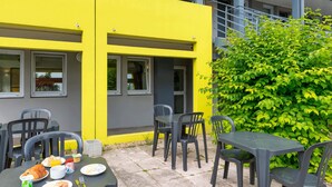 Een buitenruimte met plastic stoelen en tafels, een geel gebouw met ramen en een groene struik op de achtergrond.