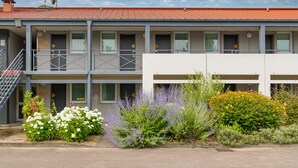 Een gebouw met twee verdiepingen, balkons, een rood dak en een tuin met verschillende planten en bloemen.