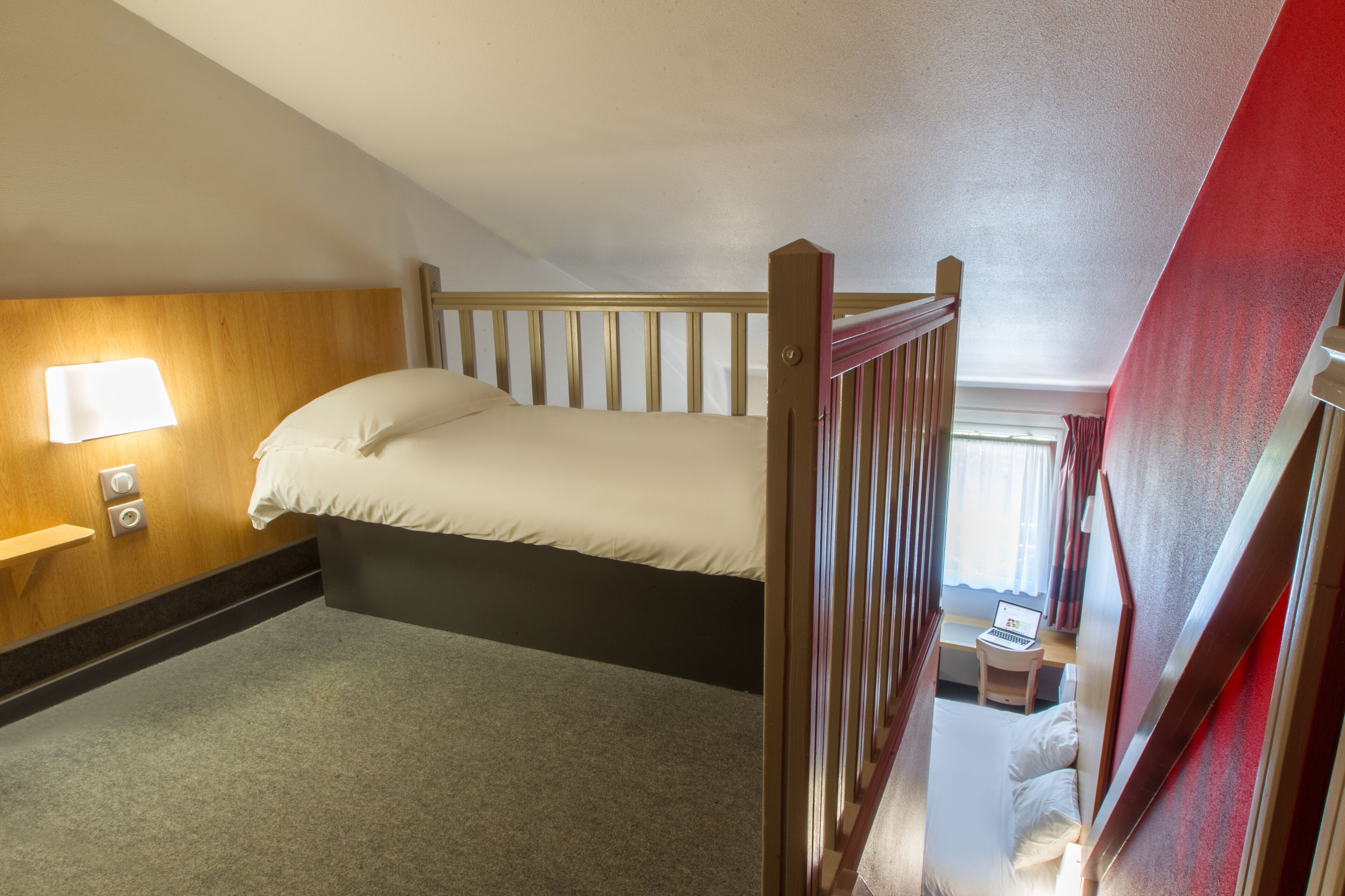 Una cama individual en una habitación con cabecera de madera, una mesita de noche con lámpara y un pequeño escritorio con un ordenador portátil.