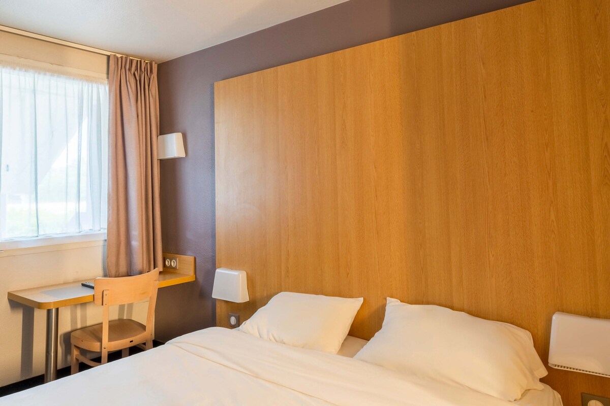 A hotel room with a bed, a desk, a chair, and a nightstand with a lamp.