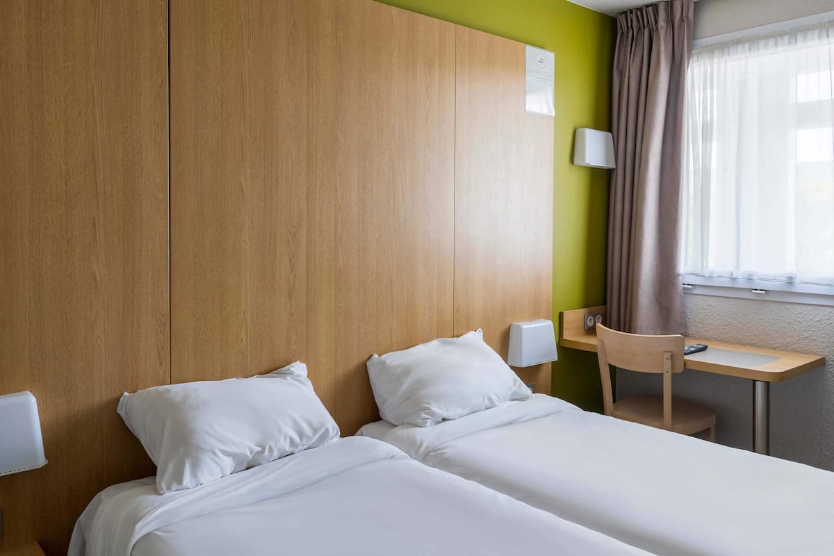Une chambre d’hôtel avec deux lits simples, une tête de lit en bois, un bureau avec une chaise, une lampe et une fenêtre avec des rideaux.
