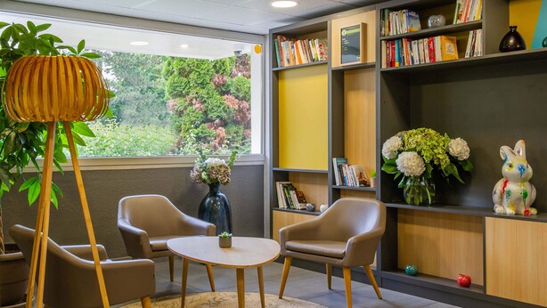 Una habitación con una mesa redonda, dos sillas, una estantería y una ventana con vista a follaje.