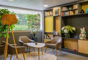 Een kamer met een ronde tafel, twee stoelen, een boekenkast en een raam met uitzicht op groen.