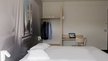 A hotel room with a bed, a desk with a computer, a chair, a wardrobe, and a window with a view of a building.
