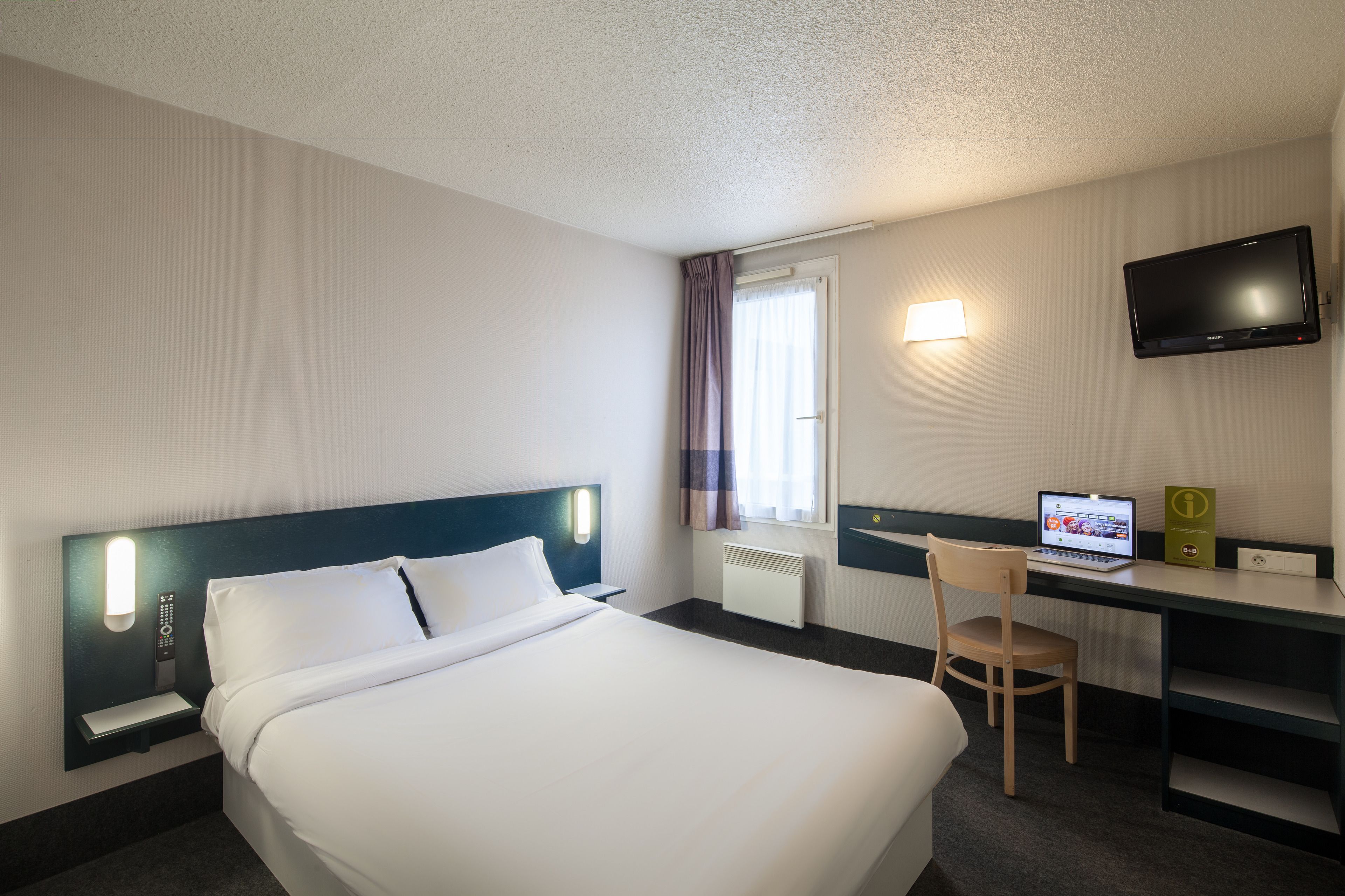 Una habitación de hotel con cama, escritorio con computadora portátil, televisión y ventana con cortinas.