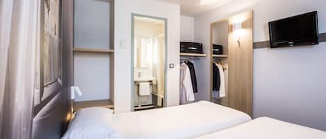 Habitación de hotel con dos camas, un televisor montado en la pared, un armario con ropa colgada y un baño visible a través de una puerta abierta.