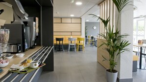 Un área de cocina moderna con refrigerador, microondas y cafetera. Hay sillas y mesas en el comedor, y se ve una planta en maceta.